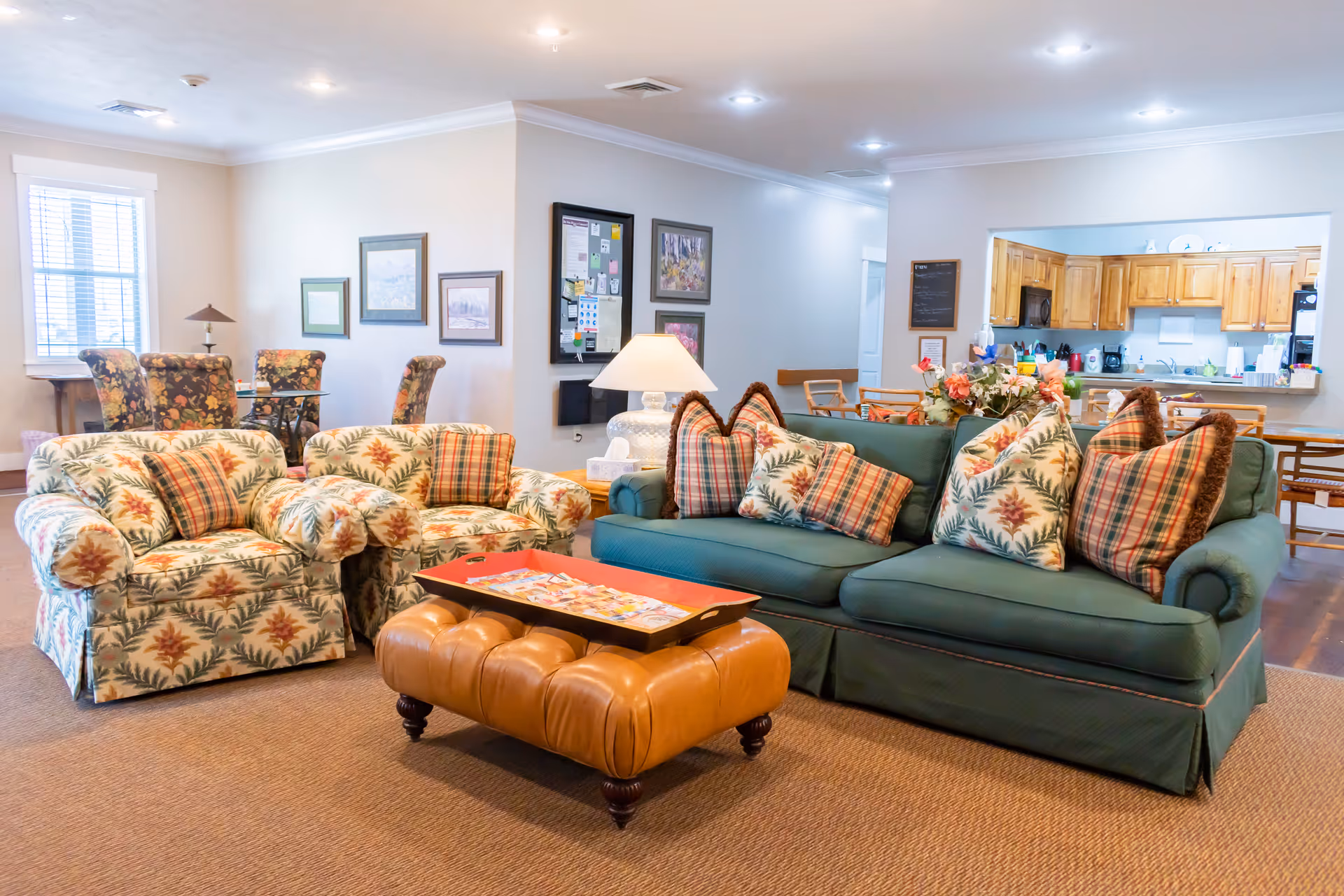 A cozy living room area in a senior living facility featuring a green sofa with patterned cushions, two matching armchairs with floral upholstery, a brown leather tufted ottoman with a red tray on top, and a glass dining table with floral upholstered chairs in the background. The room has soft lighting, framed artwork on the walls, and an open view into a kitchen with wooden cabinets.