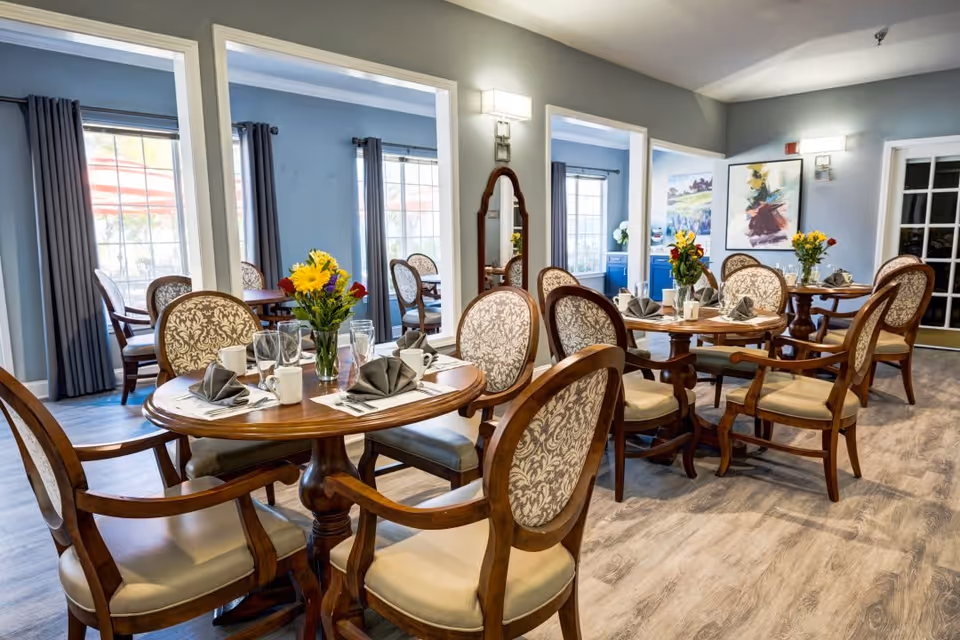 A bright and elegant dining room with wooden tables and upholstered chairs arranged neatly. Each table is set with folded gray napkins, glasses, mugs, and a vase of colorful flowers. Large windows with gray curtains allow natural light to fill the room, and decorative artwork and mirrors adorn the walls.