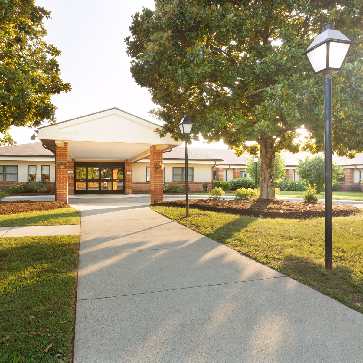 Exterior view of Heritage Hall Brookneal facility entrance with a paved walkway leading to a covered entry supported by brick columns. The area is landscaped with green grass, bushes, and large trees, and there are black lamp posts along the walkway.