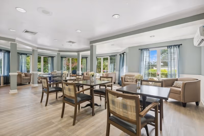 A bright and spacious common area in a senior living facility with multiple tables and chairs arranged for seating. The room features large windows with sheer blue curtains allowing natural light to fill the space. There are several comfortable armchairs and small sofas along the walls, and vases with flowers are placed on the tables. The flooring is light wood, and the walls are painted in a soft green color with white trim.