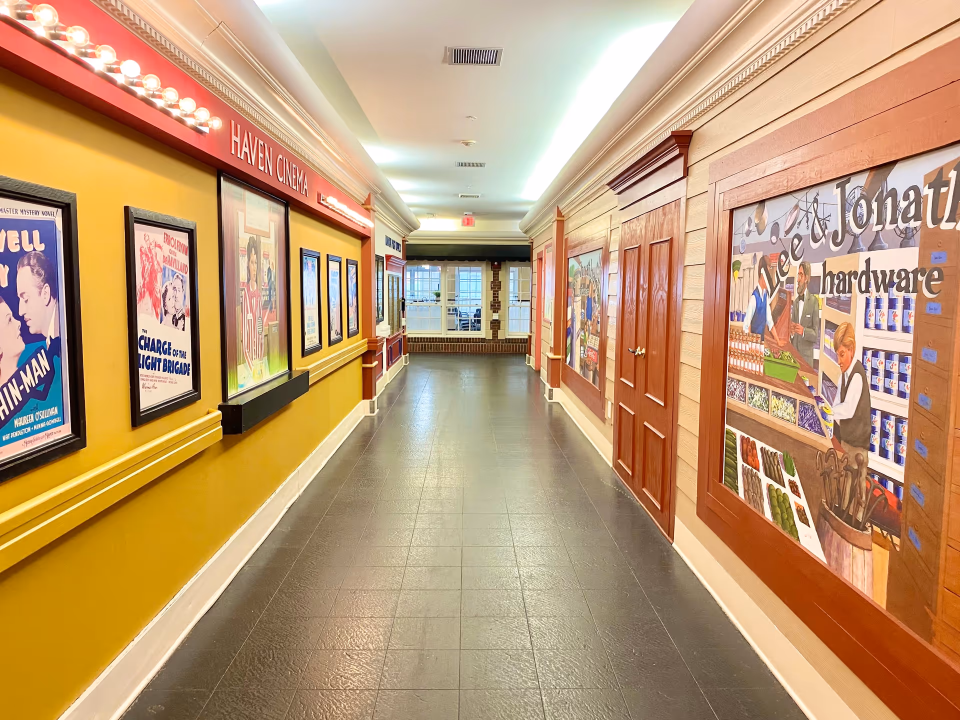 A brightly lit hallway with yellow and wooden paneled walls. On the left wall, there are framed vintage movie posters under a sign that reads 'Haven Cinema' with lights above it. The right wall features large painted murals depicting a hardware store scene. The floor is tiled in dark gray, and at the end of the hallway, there are glass doors with windows letting in natural light.