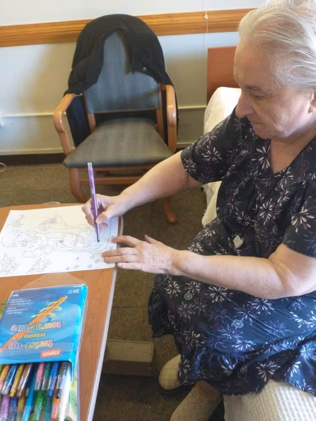 An elderly woman sitting on a bed in a room, coloring a picture with a purple crayon. There is a box of twist-up crayons on the table in front of her, and a chair with a black jacket draped over it in the background.
