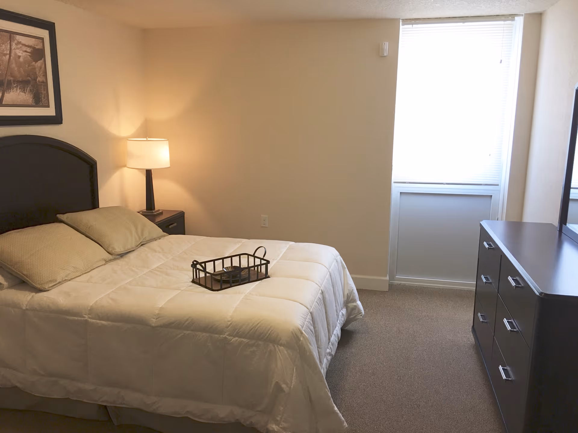 A bedroom with a neatly made bed featuring a white comforter and two pillows. A black metal tray is placed on the bed. To the left of the bed is a nightstand with a lamp, and above the bed hangs a framed picture. On the right side of the room is a dark dresser with a mirror above it. A window with blinds is on the far wall, allowing natural light into the room.
