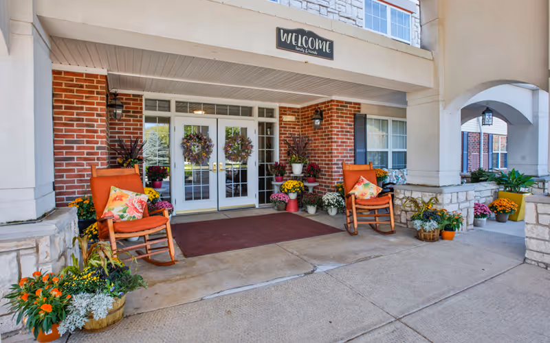 Entrance of a senior living facility with double glass doors decorated with wreaths. Two orange cushioned rocking chairs with floral pillows are placed on either side of the entrance. Various potted plants and flowers are arranged around the doorway. A 'WELCOME family & friends' sign is mounted above the doors. The building exterior features brick and stone accents with a covered walkway.
