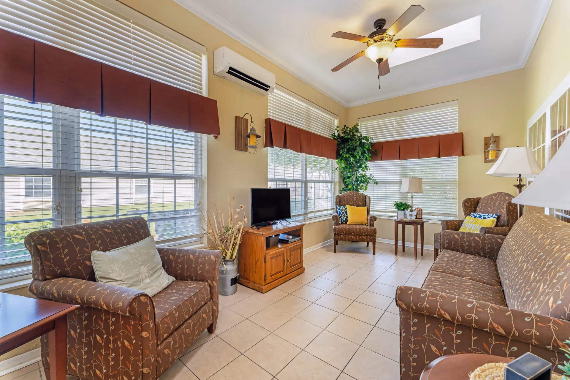 A bright living room with large windows covered by white blinds and brown valances. The room features a ceiling fan with a light, a small TV on a wooden stand, a potted plant, and several brown upholstered chairs and a sofa with leaf patterns. There are decorative pillows on the chairs and sofa, and the floor is tiled.
