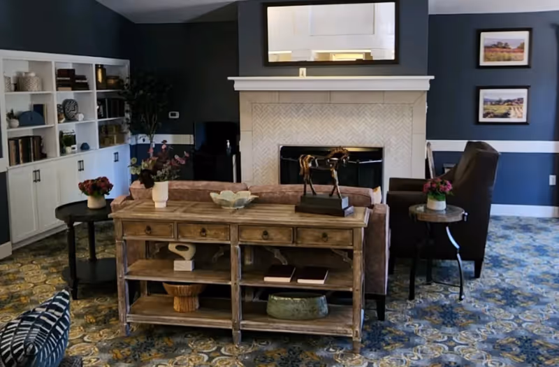 Interior living room with a fireplace and mirror, a sofa behind a wooden console table, armchairs, built-in shelving and decorative accents.