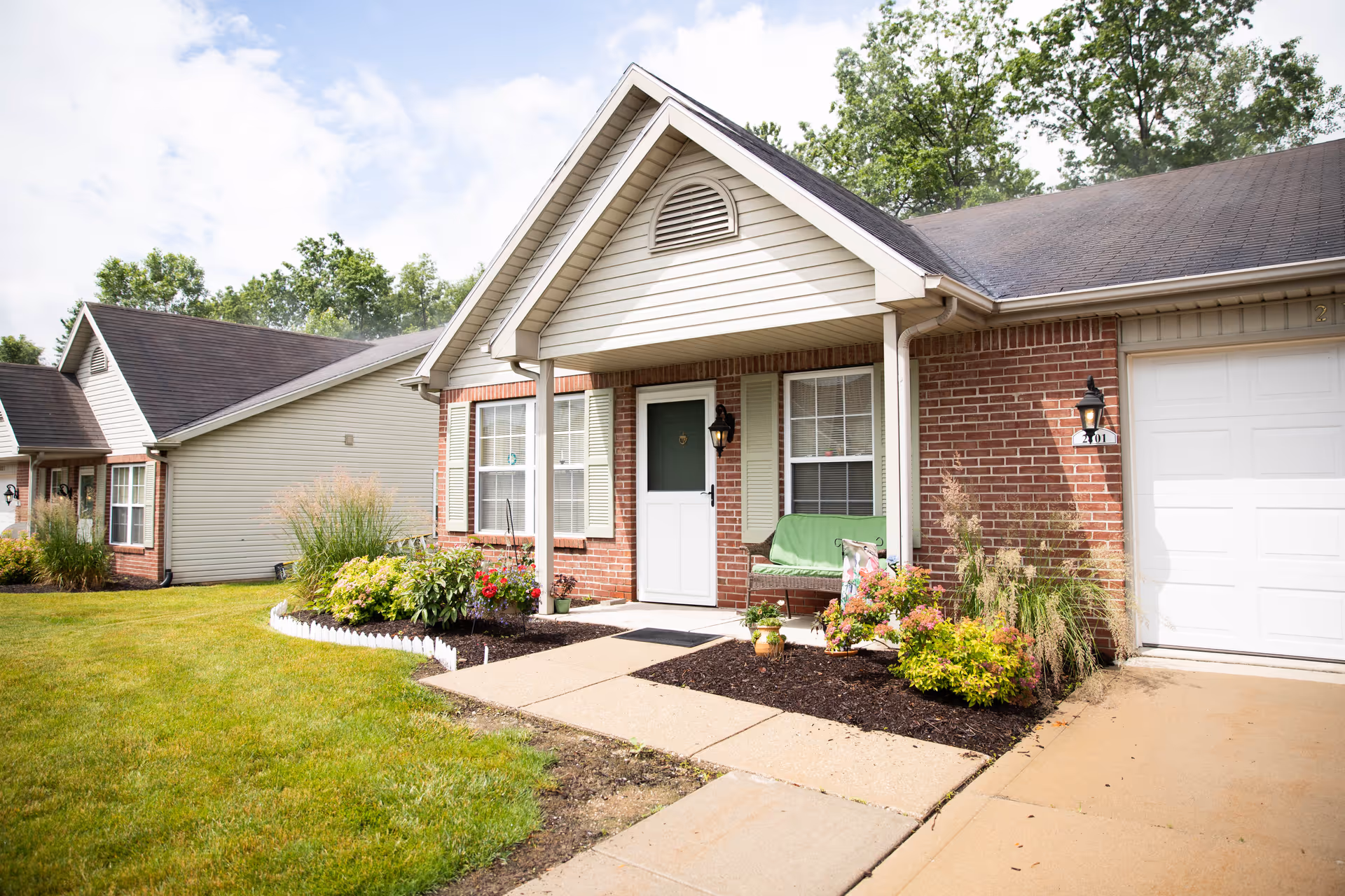 Exterior view of a single-story assisted living facility unit with a small front porch, a green bench, flower beds, and a well-maintained lawn under a partly cloudy sky.