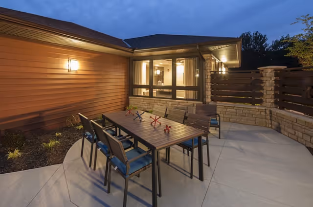 Outdoor patio at dusk with a rectangular dining table and chairs next to a building and stone privacy wall.