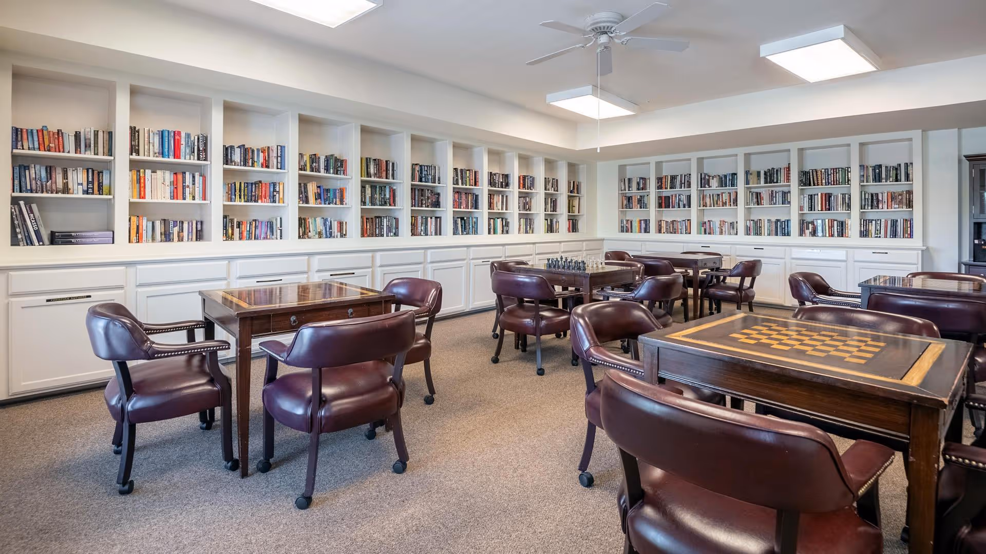 A spacious room with multiple wooden tables featuring chess boards on top, surrounded by dark leather chairs. The walls are lined with white built-in bookshelves filled with books. The room has a ceiling fan and bright overhead lighting.