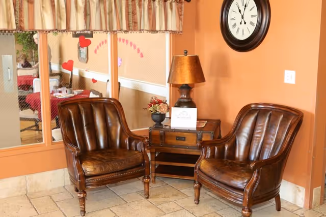 Two brown leather armchairs with wooden legs are positioned around a small wooden side table with a lamp and a flower arrangement. A large round clock hangs on the orange wall above the table. A window with a valance shows a dining area with tables and chairs decorated with red hearts.
