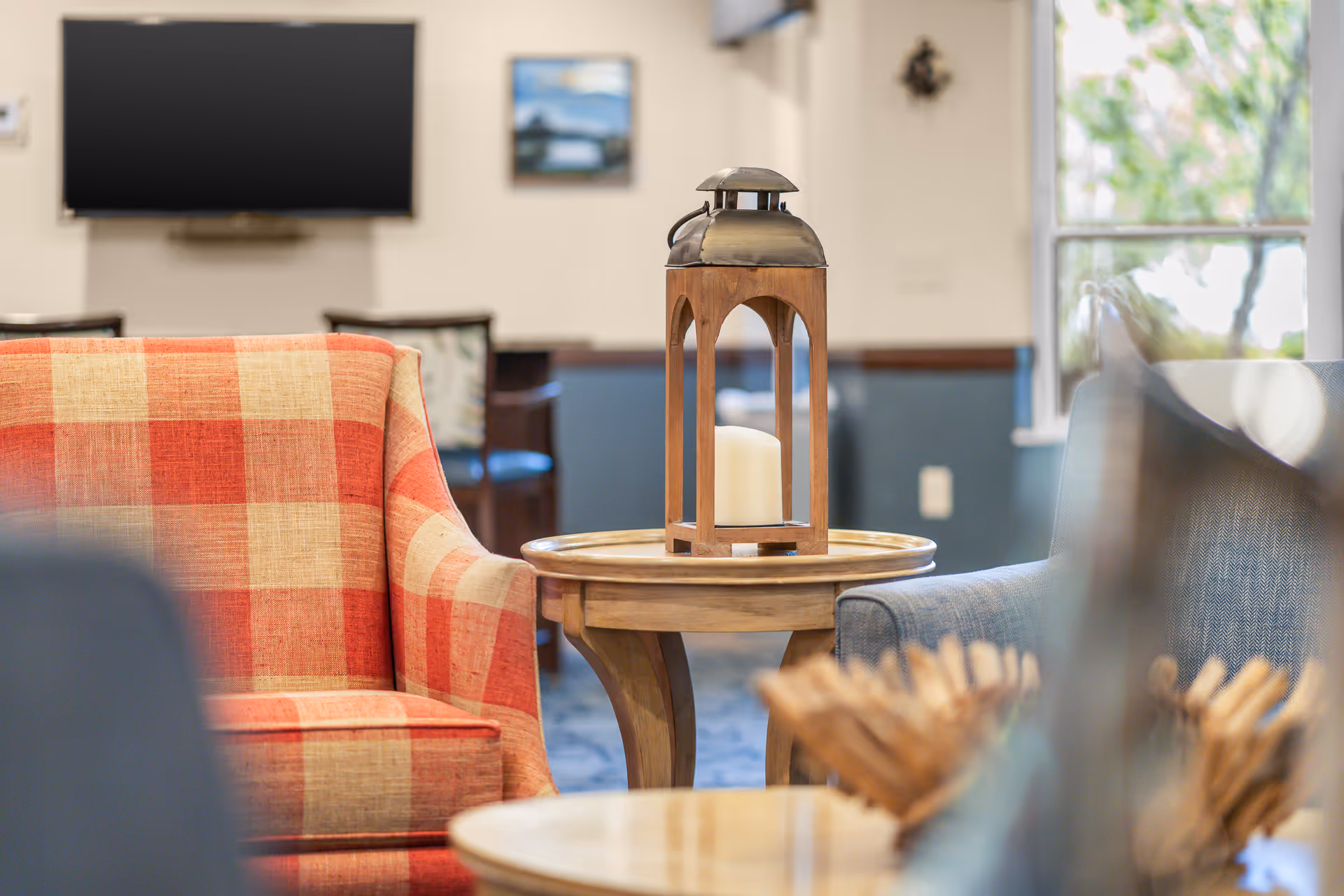 Cozy seating area in a living room with a red and beige checkered armchair, a wooden side table with a decorative lantern holding a candle, and a blue armchair. In the background, there is a wall-mounted TV and a window letting in natural light.