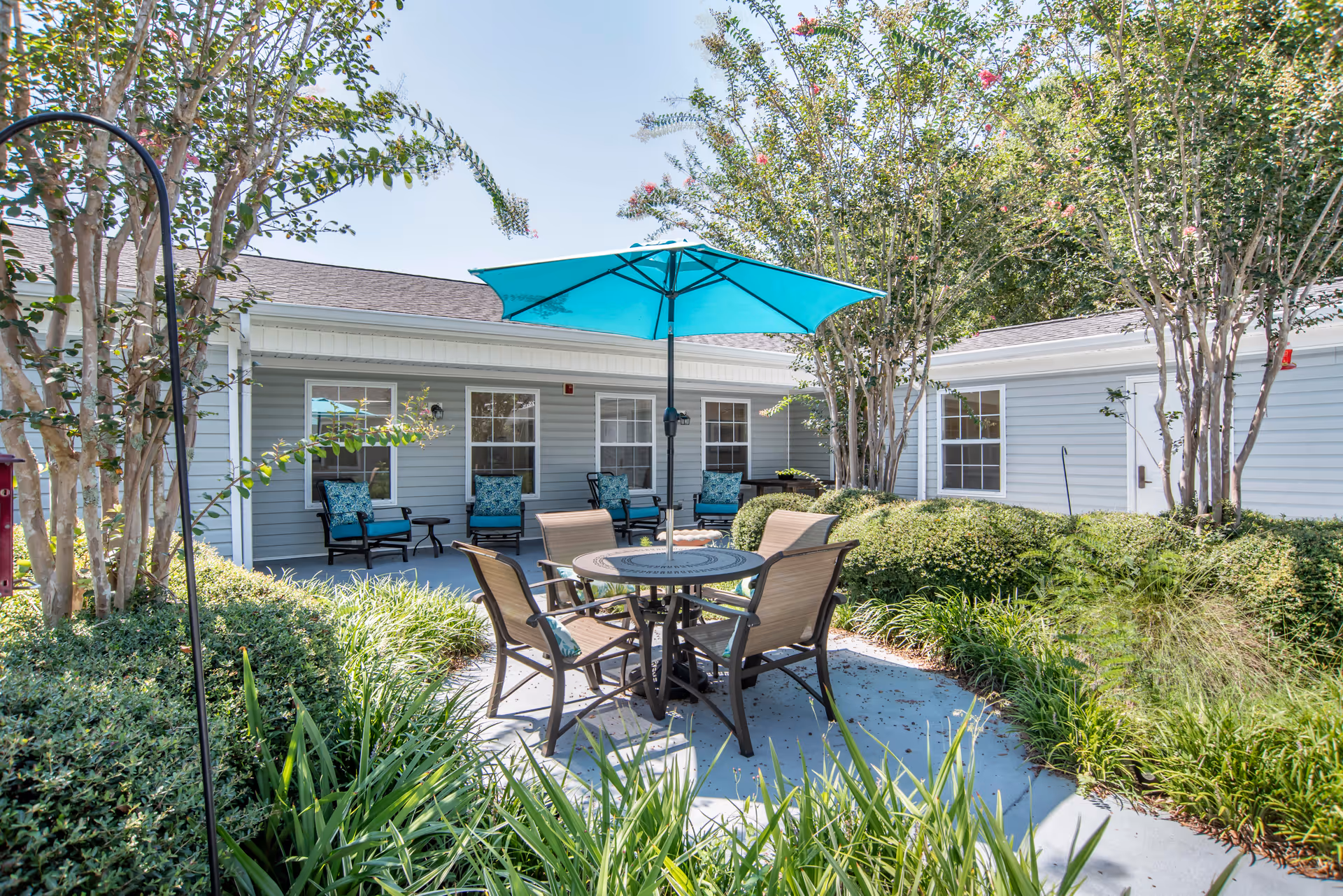 Outdoor courtyard area with a round table and four chairs under a large blue umbrella, surrounded by green bushes and trees. The background shows a single-story building with multiple windows and several cushioned chairs along the wall.