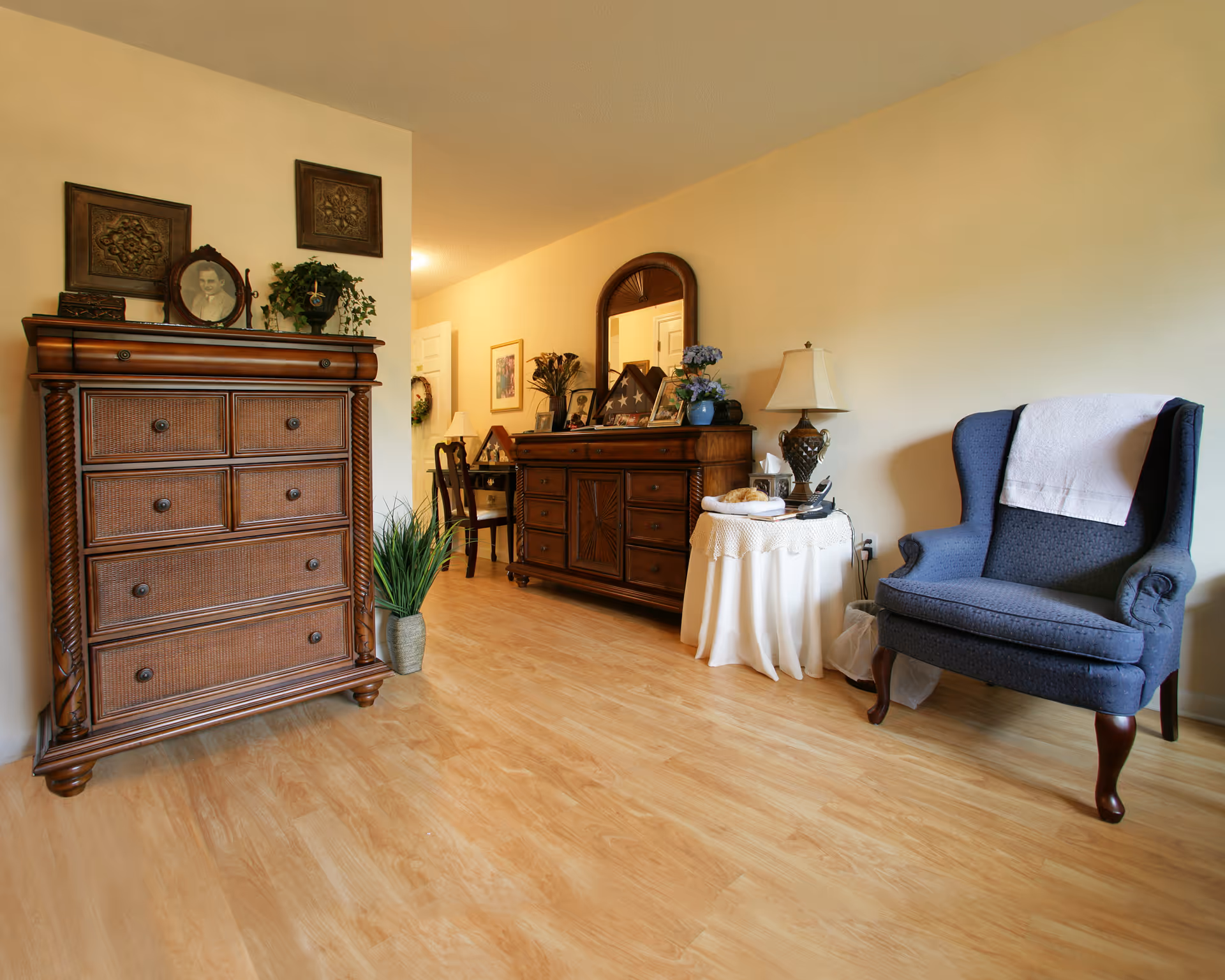 A cozy interior room with wooden flooring featuring a dark wooden chest of drawers on the left, a matching wooden sideboard with a mirror and decorative items in the center, and a blue upholstered armchair with a white cloth draped over the back on the right. A small round table with a white tablecloth, lamp, and other items is placed next to the armchair.