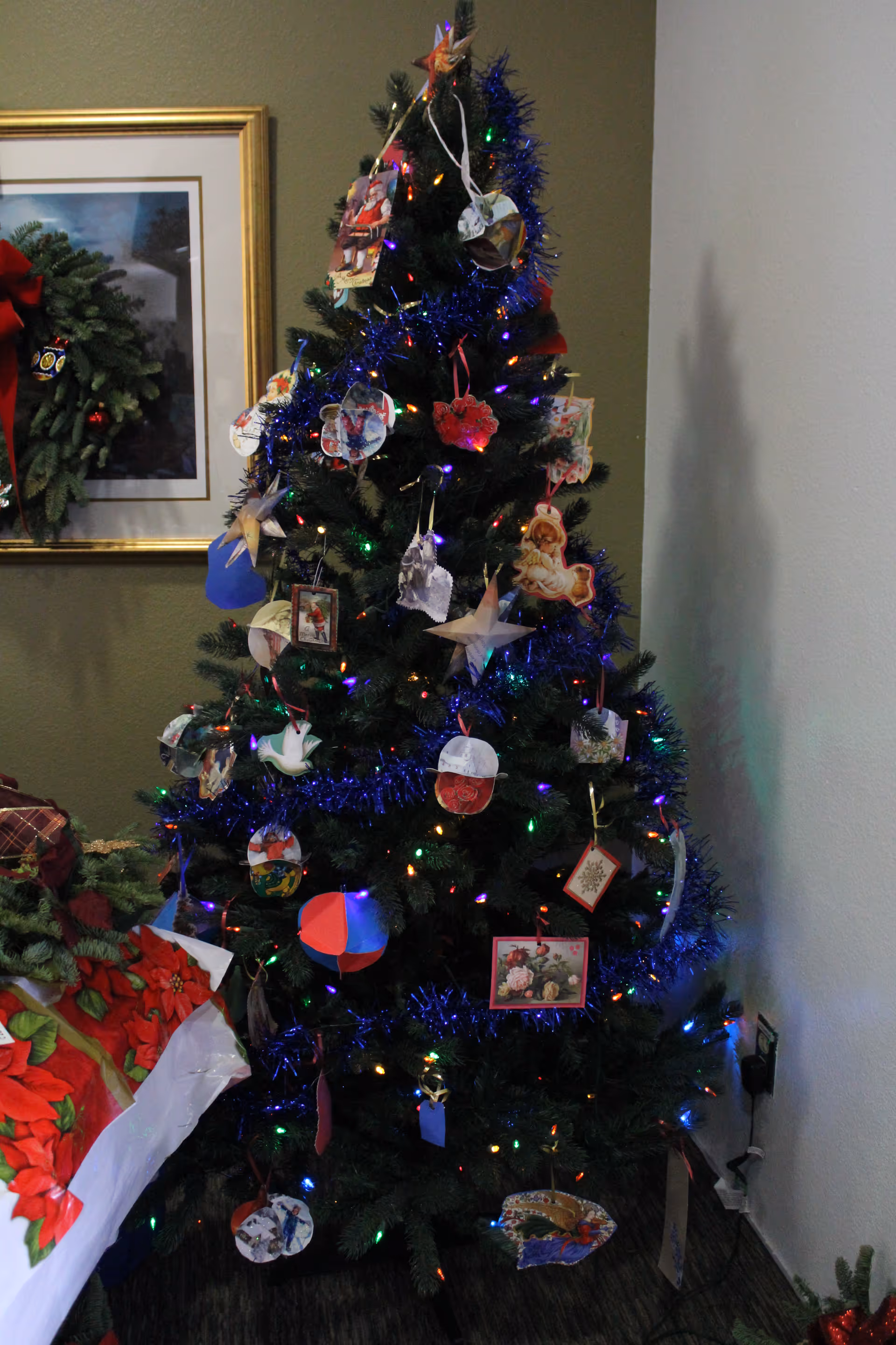 A decorated Christmas tree with colorful lights, blue tinsel, and various handmade ornaments stands in the corner of a room. To the left, there is a framed picture on the wall with a holiday wreath hanging on it, and part of a table covered with a red and green poinsettia-themed cloth is visible.