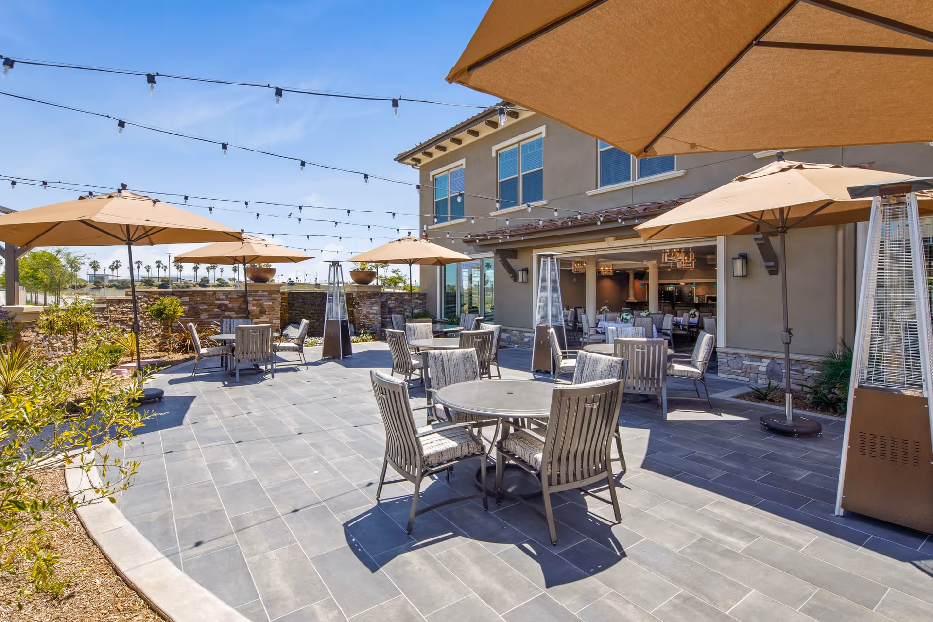Outdoor patio area with several round tables and chairs under large beige umbrellas. String lights are hung above the patio, and there are tall outdoor heaters placed around the seating area. The patio is adjacent to a building with large windows and an open entrance leading inside.