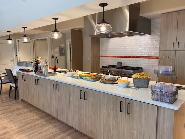 Modern kitchen area with a long island countertop featuring various food items and snacks arranged for serving. The kitchen has light wood cabinetry, a stainless steel stove with a hood, white subway tile backsplash with a red accent stripe, and pendant lights hanging above the island. The floor is wood, and there are chairs visible in the background.