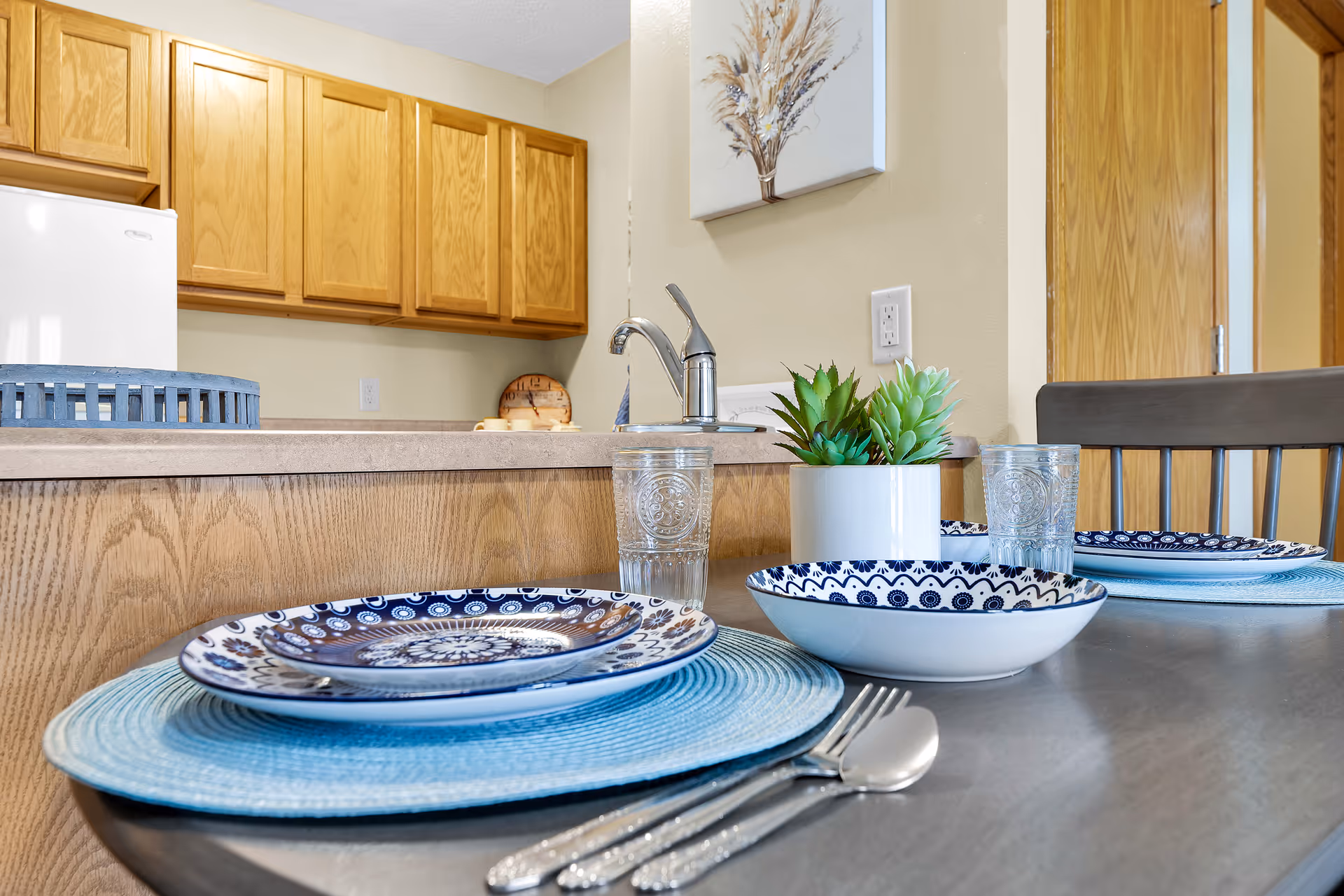 Close-up view of a dining table set with blue and white patterned plates, a bowl, clear drinking glasses, silverware, and a small white pot with green succulent plants. In the background, there is a kitchen area with wooden cabinets, a white refrigerator, a faucet, and a wall decoration featuring dried flowers.