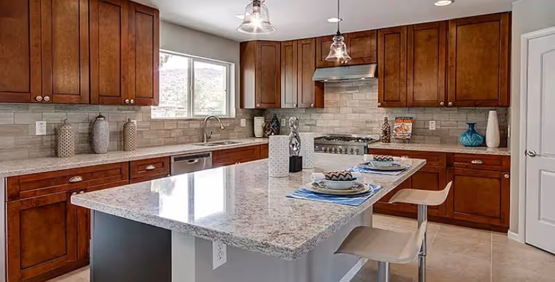 A modern kitchen with wooden cabinets, a large granite island countertop with two place settings, two pendant lights hanging above the island, a stainless steel stove with a range hood, a dishwasher, and a window letting in natural light.