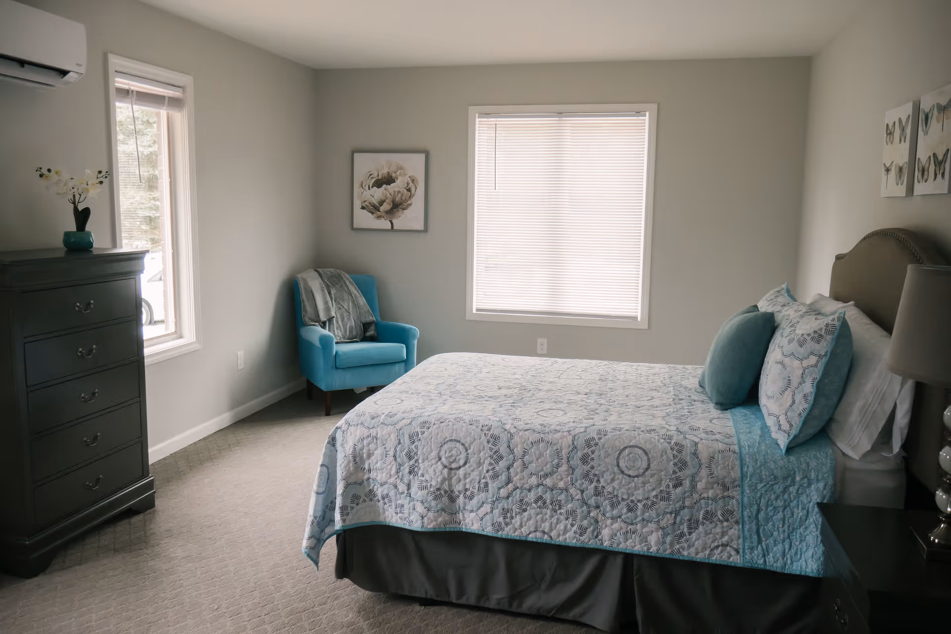 Bright bedroom with a bed dressed in teal-patterned bedding, a teal armchair, a dark dresser, and two windows.