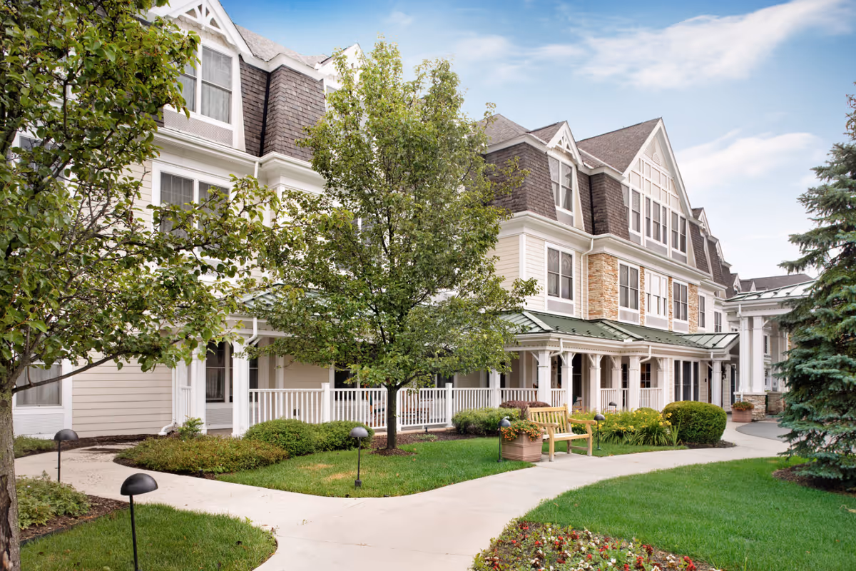Exterior view of a senior living facility named Sunrise of Bloomfield Hills, featuring a multi-story building with white and beige siding, large windows, and a green metal roof over a porch area. The foreground includes a well-maintained garden with green grass, trees, shrubs, a wooden bench, and paved walkways under a partly cloudy sky.