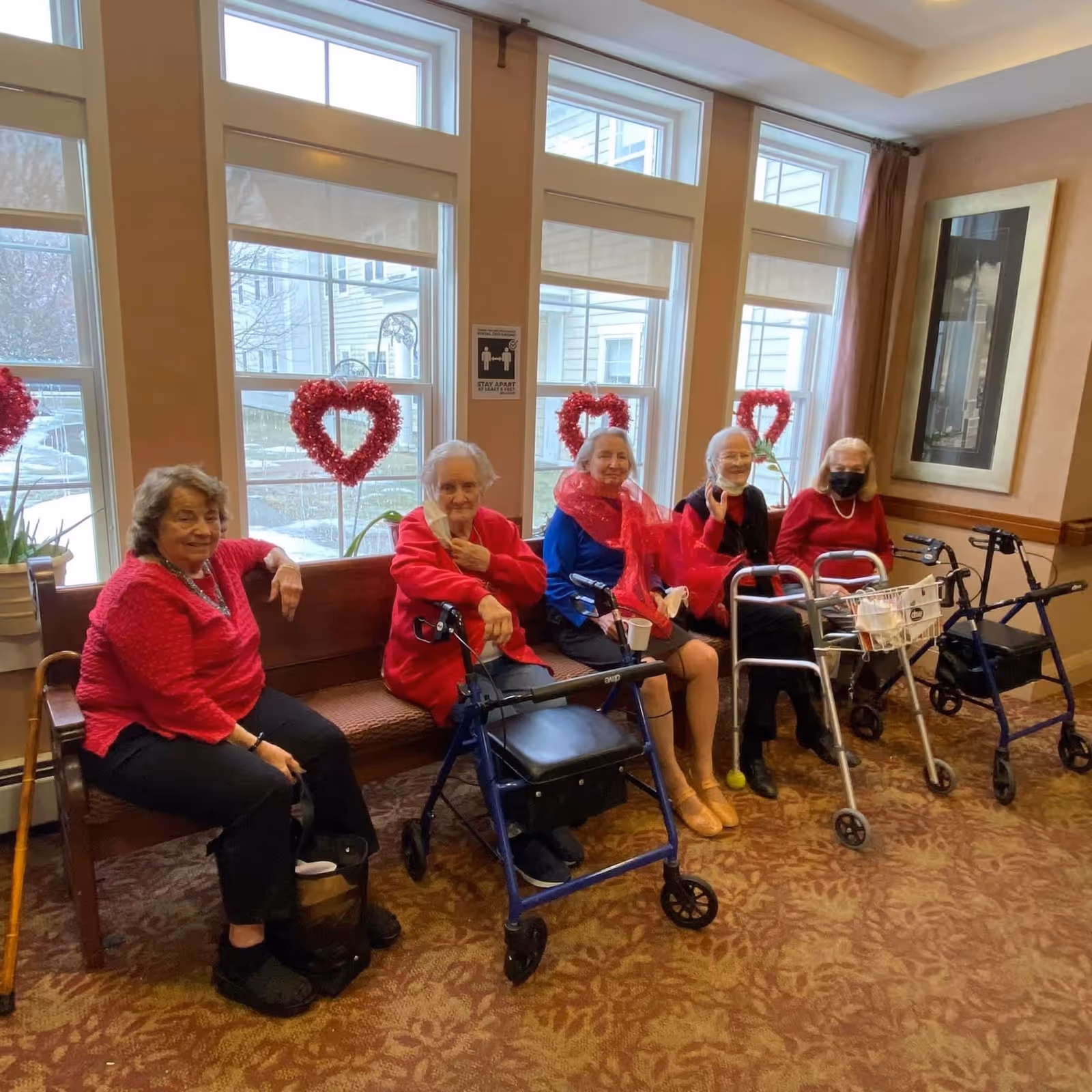 Five elderly women sitting on a wooden bench in a room with large windows decorated with red heart-shaped wreaths. Three of the women have walkers in front of them, and one woman is wearing a face mask. The room has beige walls, a patterned carpet, and a framed picture on the wall.