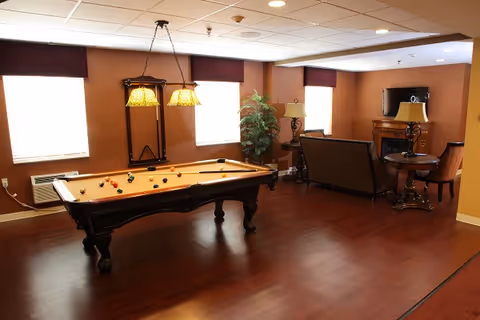 A recreational room with a pool table in the center, two hanging lights above it, and a seating area with a sofa, chairs, a TV mounted above a fireplace, and a side table with a lamp. The room has wooden flooring, brown walls, and three windows with dark valances.