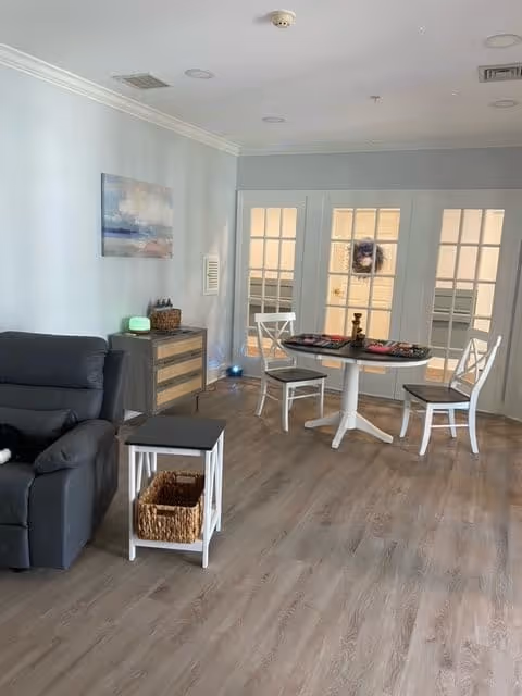 A cozy interior room with light gray walls and wood flooring. There is a dark gray recliner chair on the left, a small white side table with a wicker basket underneath, and a wooden dresser with a green diffuser and basket on top. In the background, there is a round white table with three white chairs around it, and French doors with glass panes. A painting hangs on the wall above the dresser.