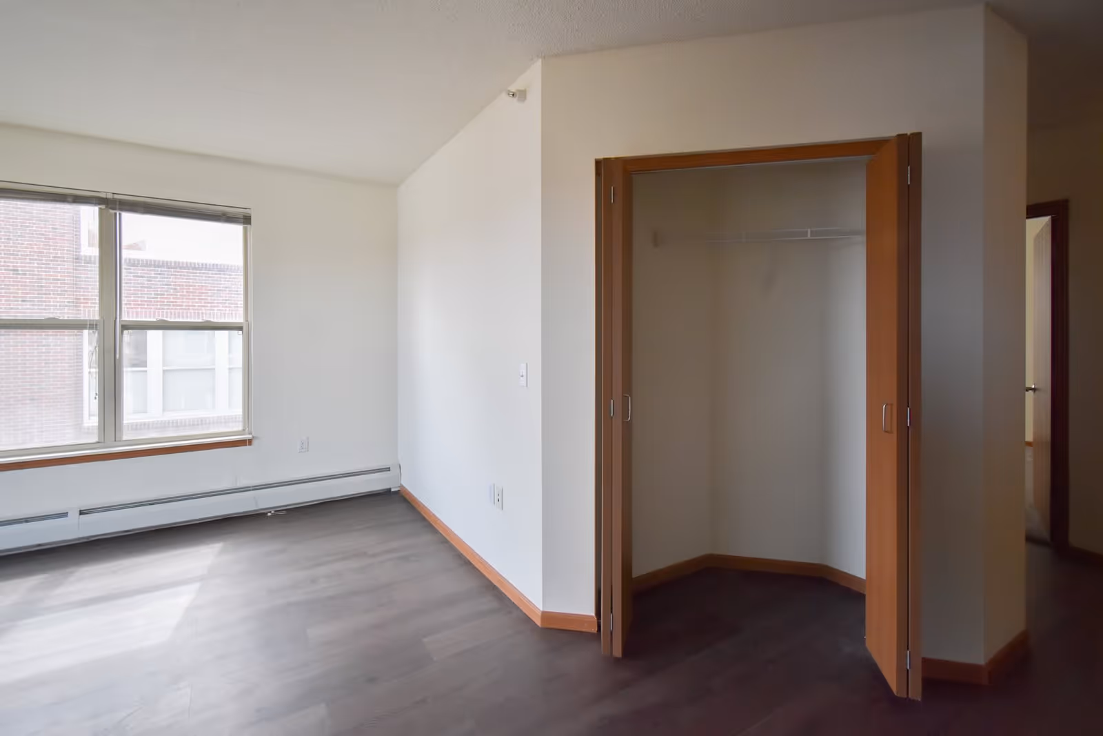 Empty room in Lyons Court Senior Apartments with dark wood flooring, white walls, a large window letting in natural light, and an open closet with wooden bi-fold doors.