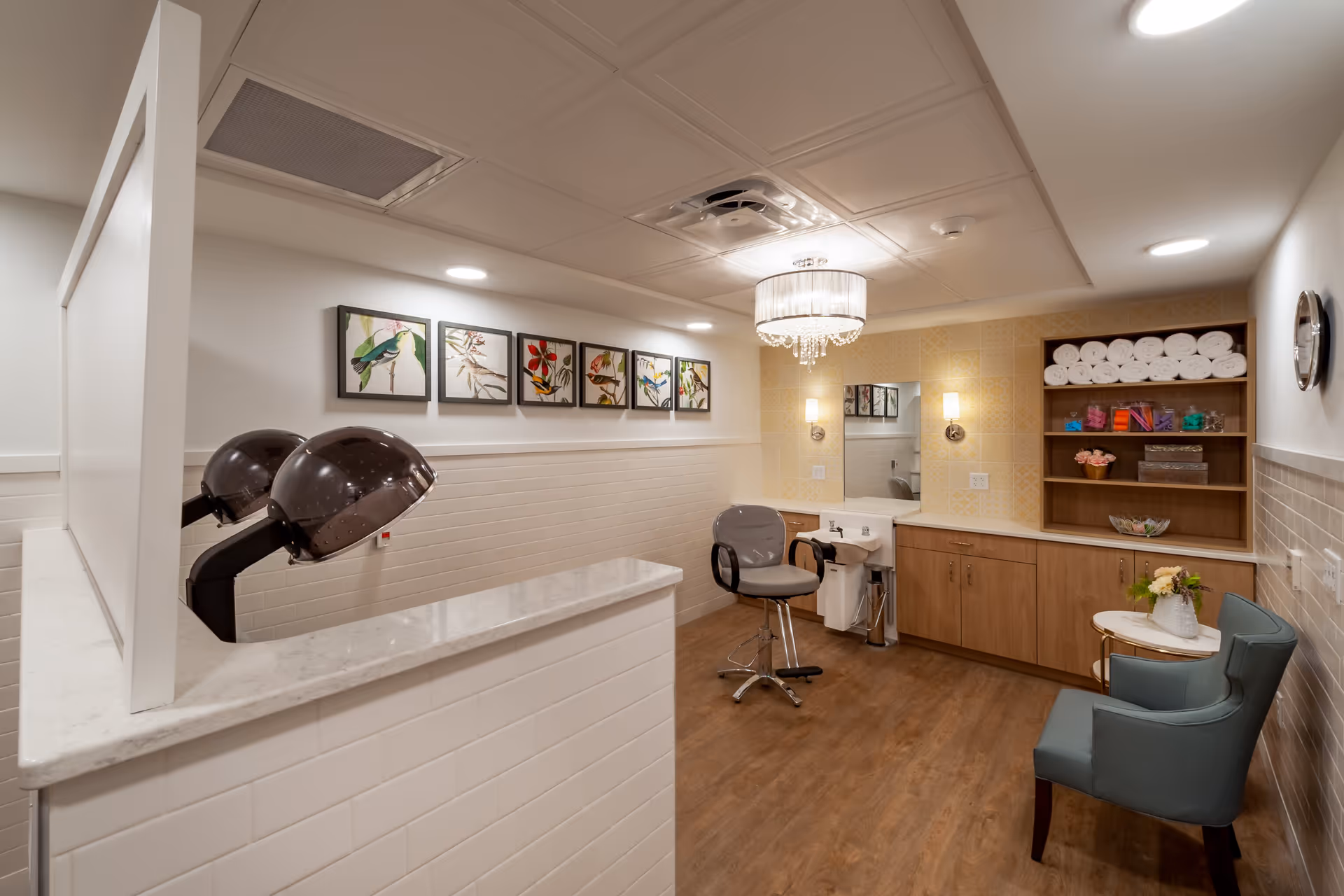 A well-lit salon area in a senior living facility with two black hair dryers mounted on a white tiled counter, a salon chair in front of a sink, wooden cabinets with shelves stocked with rolled towels and hair products, a small round table with a flower vase, and two blue upholstered chairs. The walls are decorated with framed bird and flower artwork, and a chandelier hangs from the ceiling.