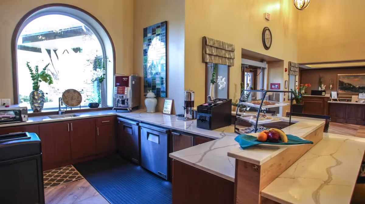 Interior view of a kitchen area in a senior living facility with a large arched window, countertop with a coffee machine, a display case with pastries, and a plate of fruit on a marble counter. The background shows a reception area with flowers and a painting on the wall.