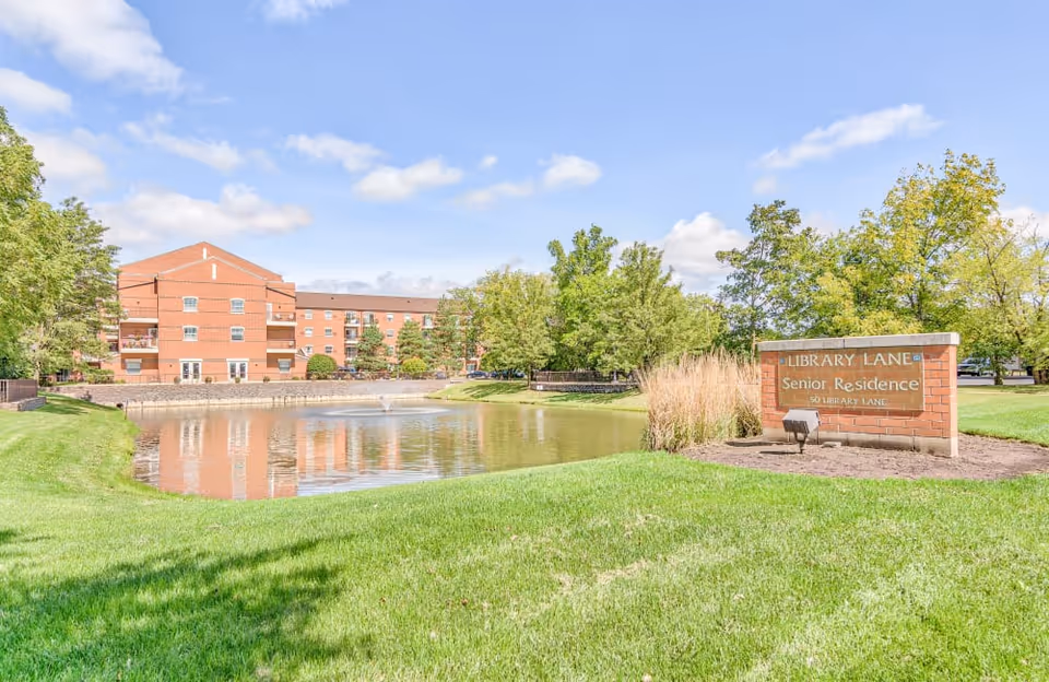 Exterior view of Library Lane Senior Residence with a brick building, pond, and a sign on the lawn.