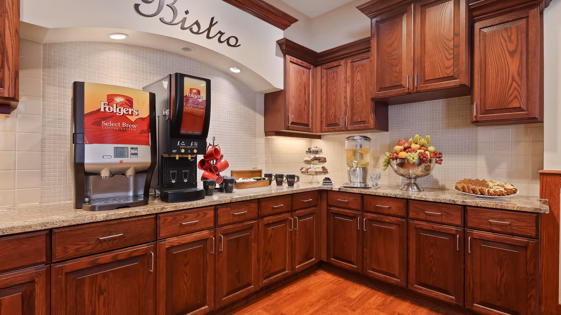 A cozy bistro area with wooden cabinets and granite countertops featuring a Folgers Select Brew coffee machine, a Folgers Cappuccino machine, red coffee mugs hanging on hooks, a glass dispenser with water and lemon slices, a tiered tray with muffins, a large bowl of assorted fresh fruit, and a plate of sliced bread or cake.