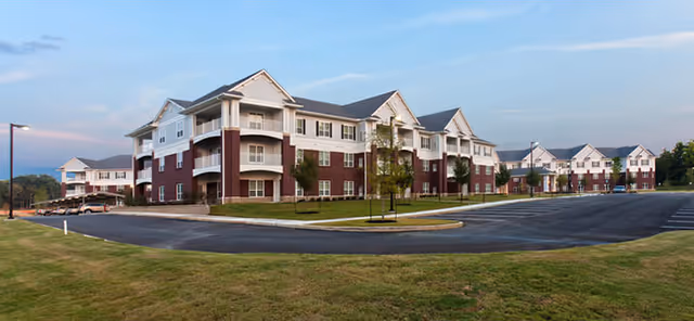 Exterior front view of a three-story assisted living building with red brick and white siding, surrounded by a parking lot and lawn.