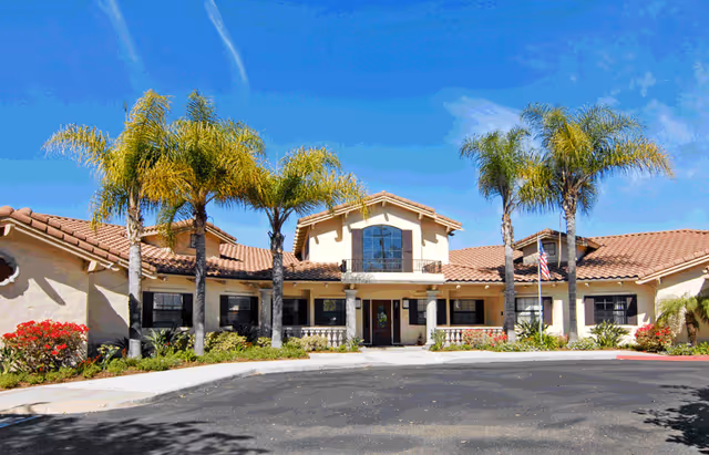 Front exterior of a Mediterranean-style memory care building with palm trees lining the entrance and a circular driveway.