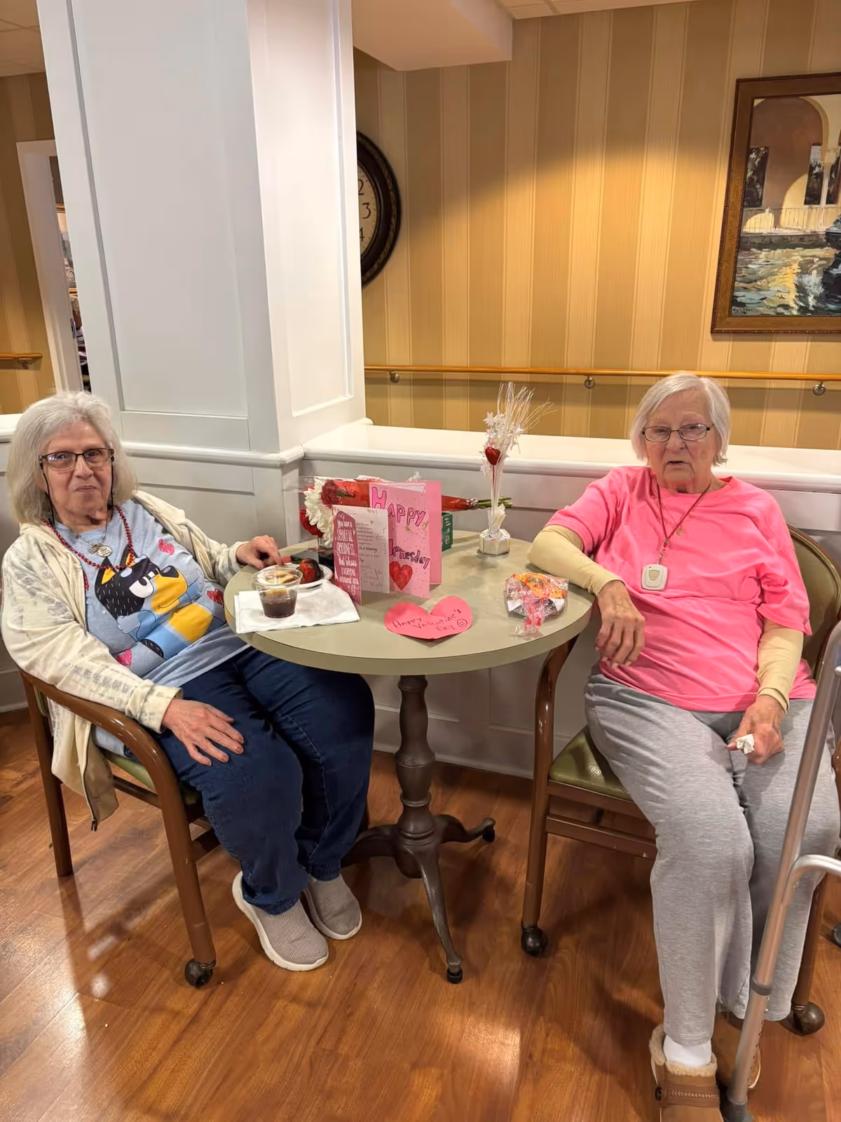 Two elderly women sitting at a small round table in a common area. The woman on the left is wearing glasses, a light jacket, and a blue shirt with a cartoon character. The woman on the right is wearing glasses, a pink shirt, and gray pants, with a walker beside her. On the table are Valentine-themed cards, a small bouquet, and some snacks. The background shows a beige wall with a framed painting and a clock partially visible.