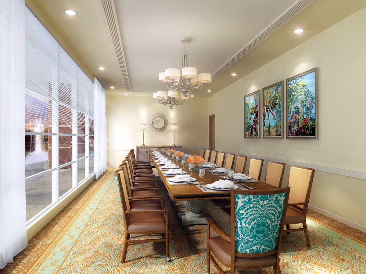 A long dining table set for a meal with white plates, napkins, and glassware in a well-lit dining room. The room features large windows with white curtains on the left side, a patterned carpet, and three framed tropical-themed paintings on the right wall. There are multiple wooden chairs with beige cushions around the table, and two lamps with a round mirror on the far wall under a chandelier.