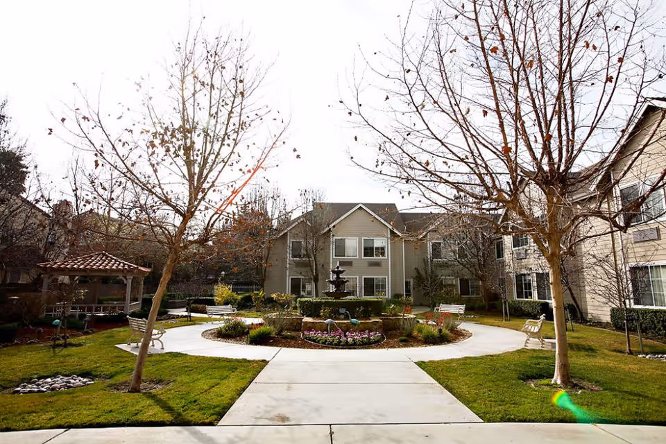 Outdoor courtyard area of a senior living facility with a circular walkway surrounding a central fountain and flower bed. There are leafless trees, benches along the path, and a gazebo on the left side. The building with multiple windows is visible in the background.