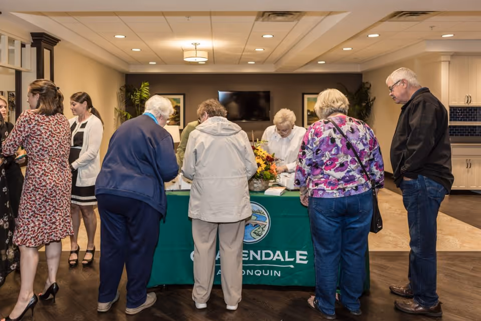 A group of elderly people and staff gathered around a green table with a Clarendale of Algonquin banner in a well-lit interior space. The table has a flower arrangement and some papers, and the background shows a television mounted on the wall and framed pictures.