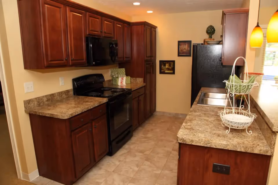 A kitchen with dark wood cabinets, a black stove and microwave, a black refrigerator, and beige countertops. The floor is tiled, and there are two yellow pendant lights hanging over the counter with a white decorative basket on it. Two framed pictures hang on the wall near the refrigerator.