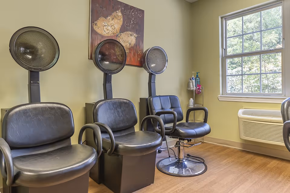 Interior view of a hair salon area with three black salon chairs, each with a hair dryer hood above. There is a window on the right side letting in natural light, a wall-mounted air conditioning unit below the window, and a decorative painting on the wall behind the chairs. Various hair care products are placed on a small stand near the window.