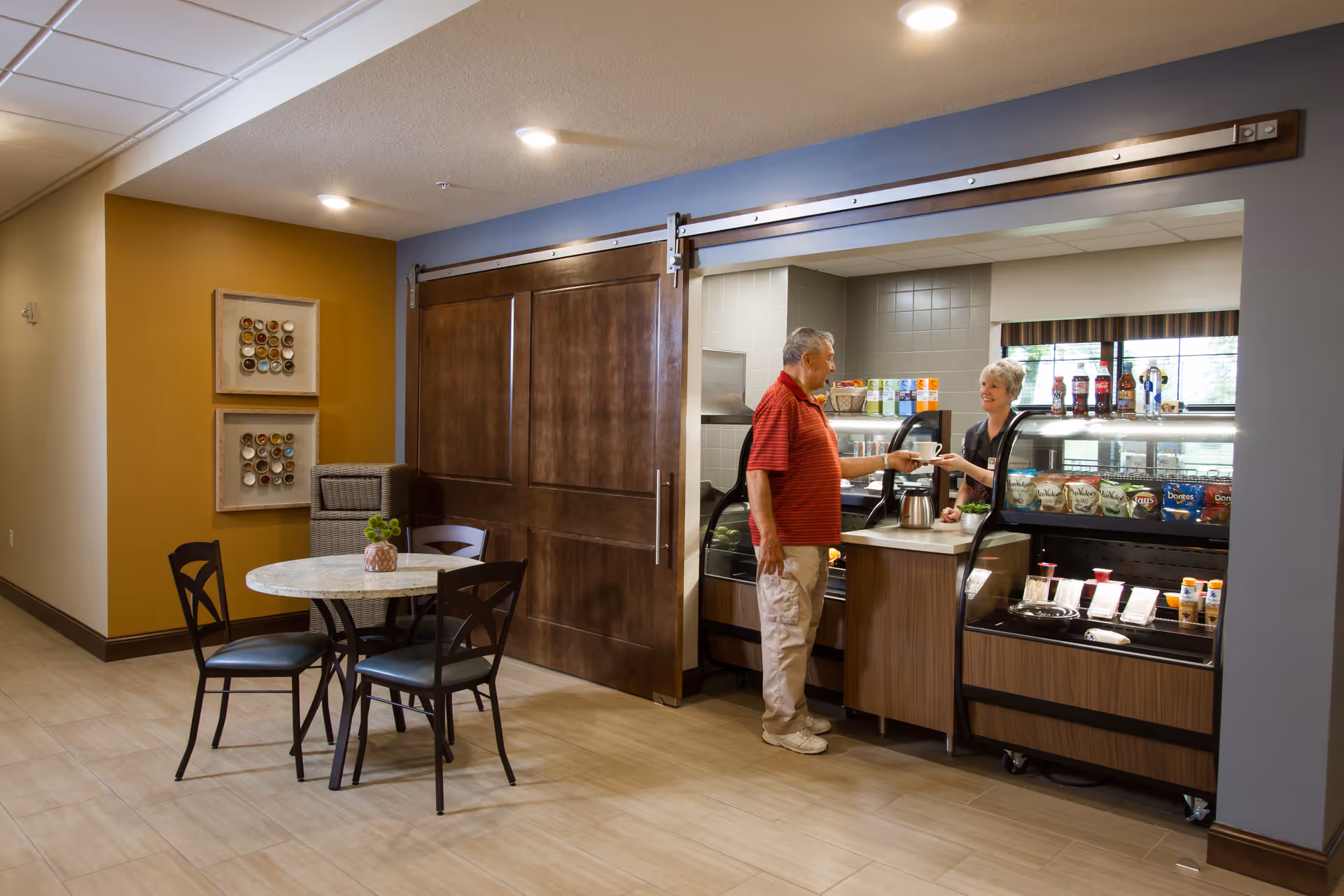 A small indoor cafe area with a table and chairs where a man is receiving a drink from a staff member at a service counter.