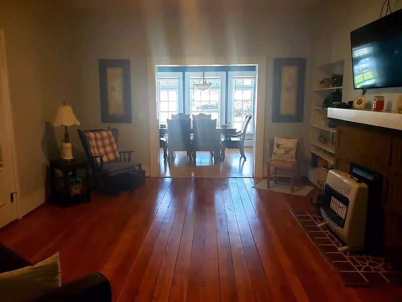Cozy living room with polished wood floors, armchairs and a fireplace opening into a sunlit dining area through a wide doorway.