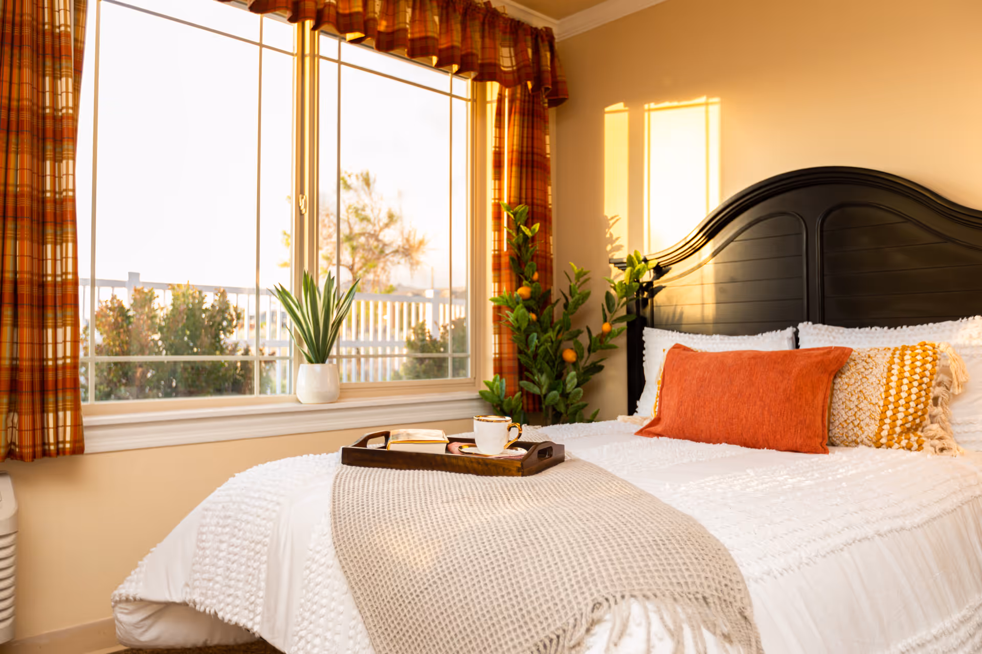 A cozy bedroom with a large window featuring plaid curtains. The bed has a dark wooden headboard and is made with white bedding, an orange rectangular pillow, and a textured yellow and white pillow. A beige knitted throw blanket is draped over the bed with a wooden tray holding a cup and a book. There is a potted plant on the windowsill and a larger plant with orange fruit beside the bed. Warm sunlight is streaming through the window, casting shadows on the wall.