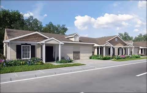 Exterior view of a single-story residential building with beige siding, dark shutters, and a brown shingled roof under a partly cloudy sky. The building has multiple entrances with small porches and a driveway in front.