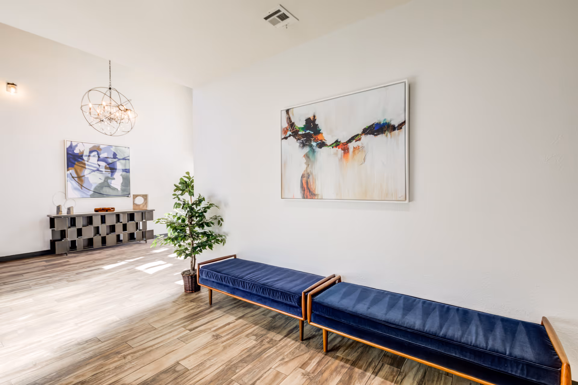A bright, modern interior space with two blue cushioned benches along a white wall. Above the benches is an abstract painting with splashes of color. On the left side, there is a decorative console table with abstract art above it and a spherical chandelier hanging from the ceiling. A potted plant is placed near the benches on a wooden floor.
