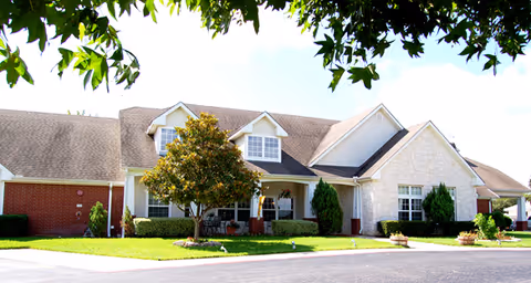 Exterior view of a single-story residential building with a combination of brick and light-colored siding. The building has multiple windows, a covered porch area with seating, and is surrounded by a well-maintained lawn with shrubs and a tree in front. The sky is clear with some tree branches visible at the top of the image.