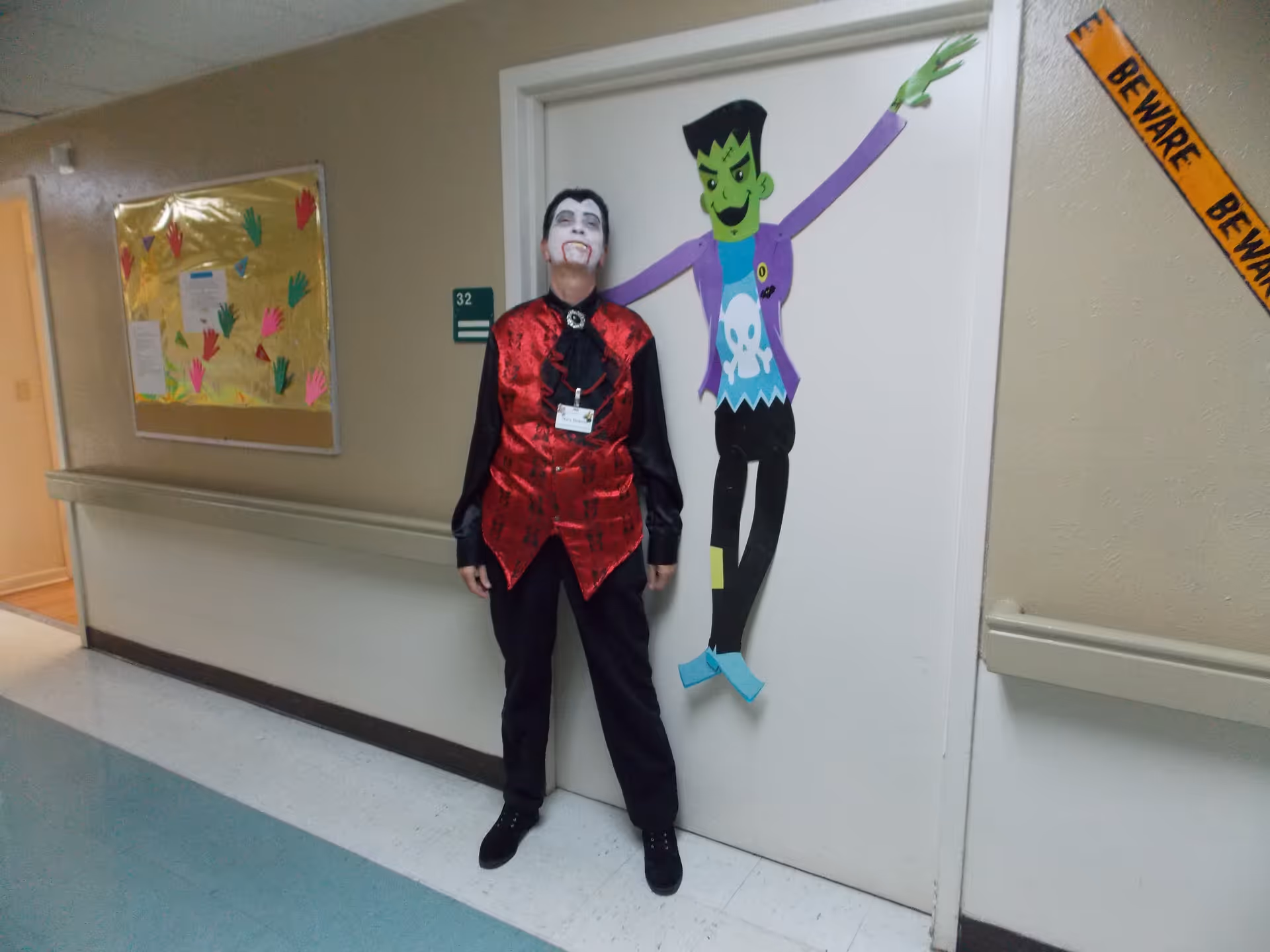 A person dressed as a vampire stands in a nursing home hallway next to a paper Frankenstein decoration on a door.