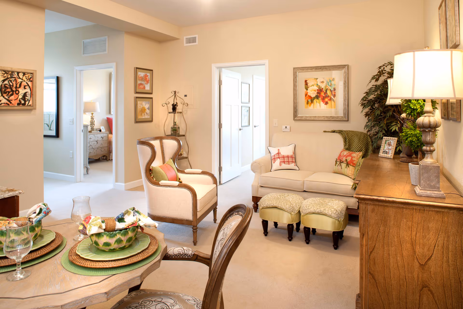 A cozy living room and dining area in an assisted living facility. The living room features a beige loveseat with decorative pillows, two small ottomans, a wooden sideboard with a lamp and framed photos, and a large plant. The dining area has a round wooden table set with green plates, napkins, and glasses. The walls are decorated with framed floral artwork, and two doorways lead to other rooms.