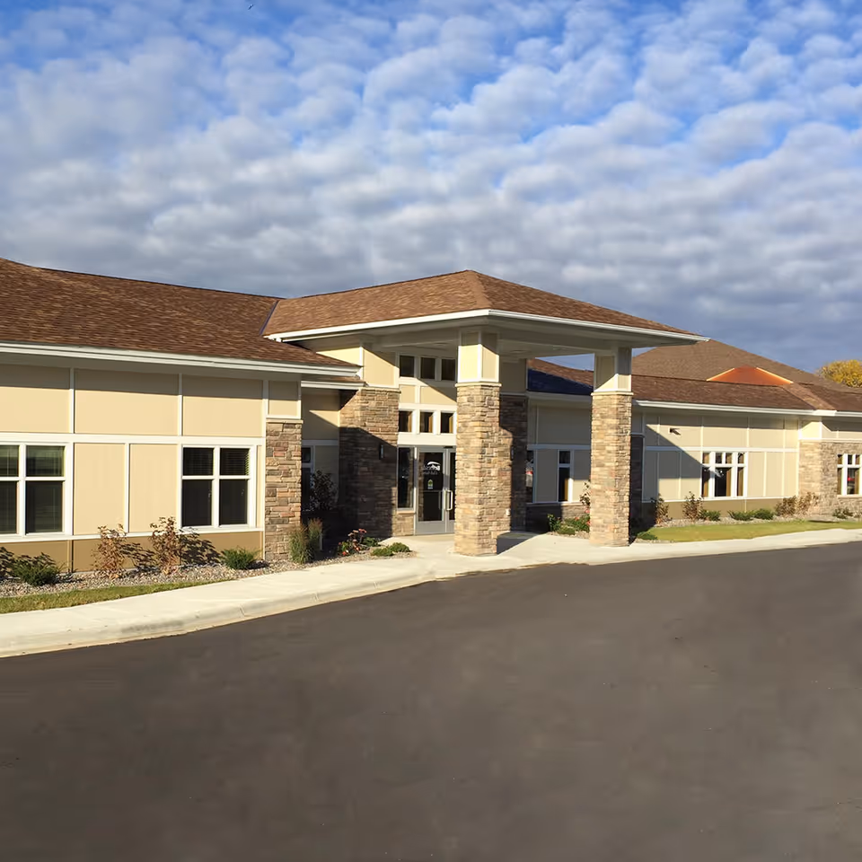 Front exterior of a single-story building with a covered entrance supported by stone pillars and a paved driveway.