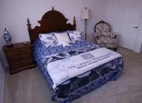 A bedroom with a wooden headboard bed covered in blue and white bedding, including pillows and a blanket that reads 'The Legacy of Anderson Senior Living Community'. To the left of the bed is a wooden nightstand with a blue and white vase and a small item on it. To the right is a floral upholstered armchair and a floor lamp. The room has light-colored carpet and white walls with a closed white door in the background.